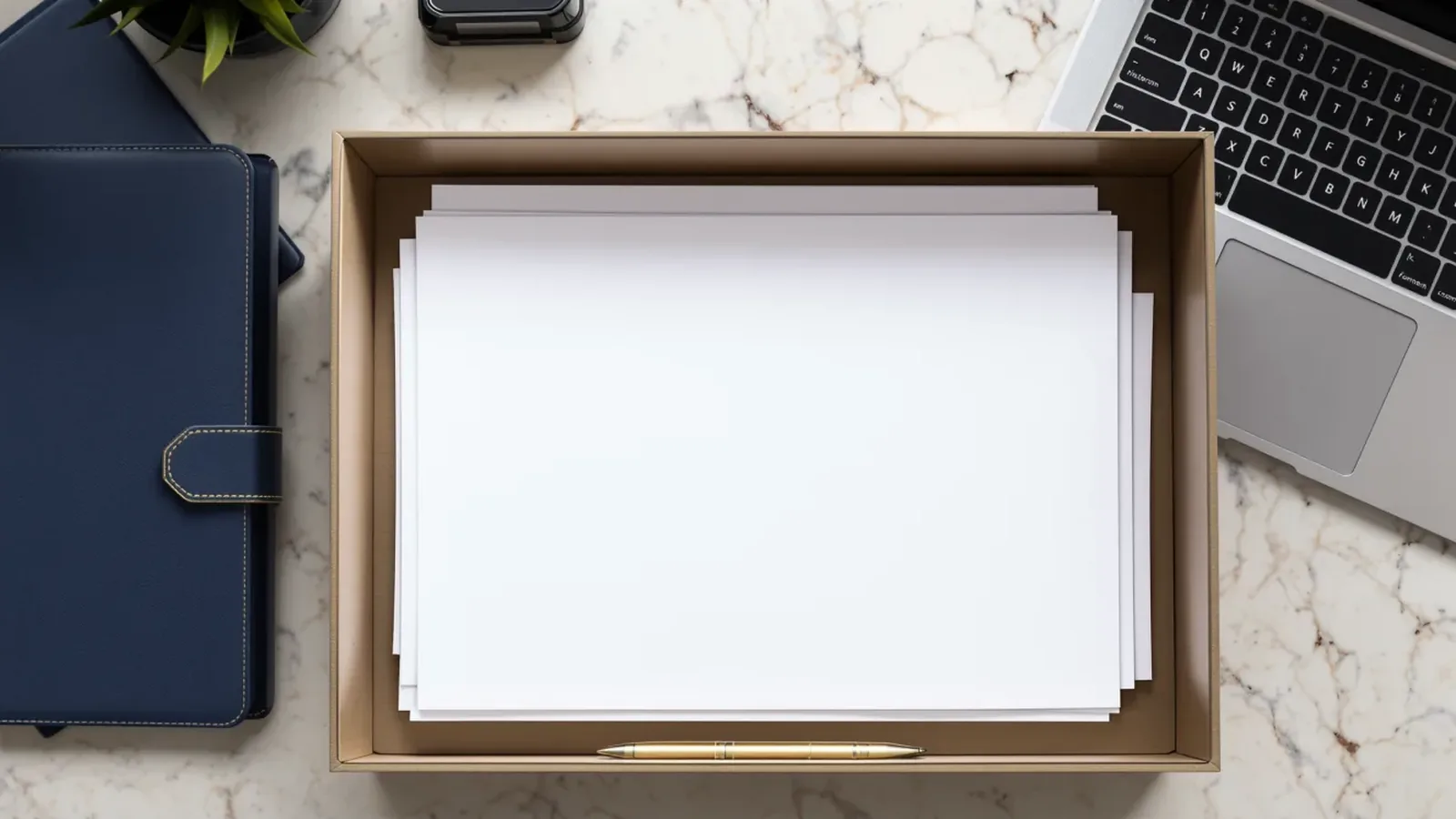 Top-down view of a marble desk with a kraft document tray holding a stack of blank white paper and a brass pen, a navy leather folio on the left, and an open laptop on the right